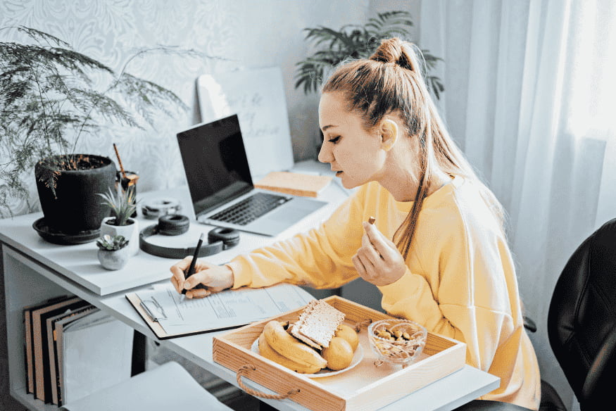 Donna che compila un modulo fiscale mentre lavora da casa con laptop e snack sulla scrivania.