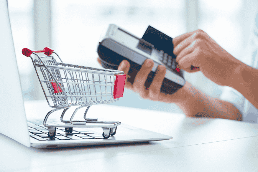 Mano che usa una carta su un POS accanto a un carrello della spesa su laptop, immagine legata ad autenticazione forte, e-commerce e carta rifiutata nei pagamenti online.